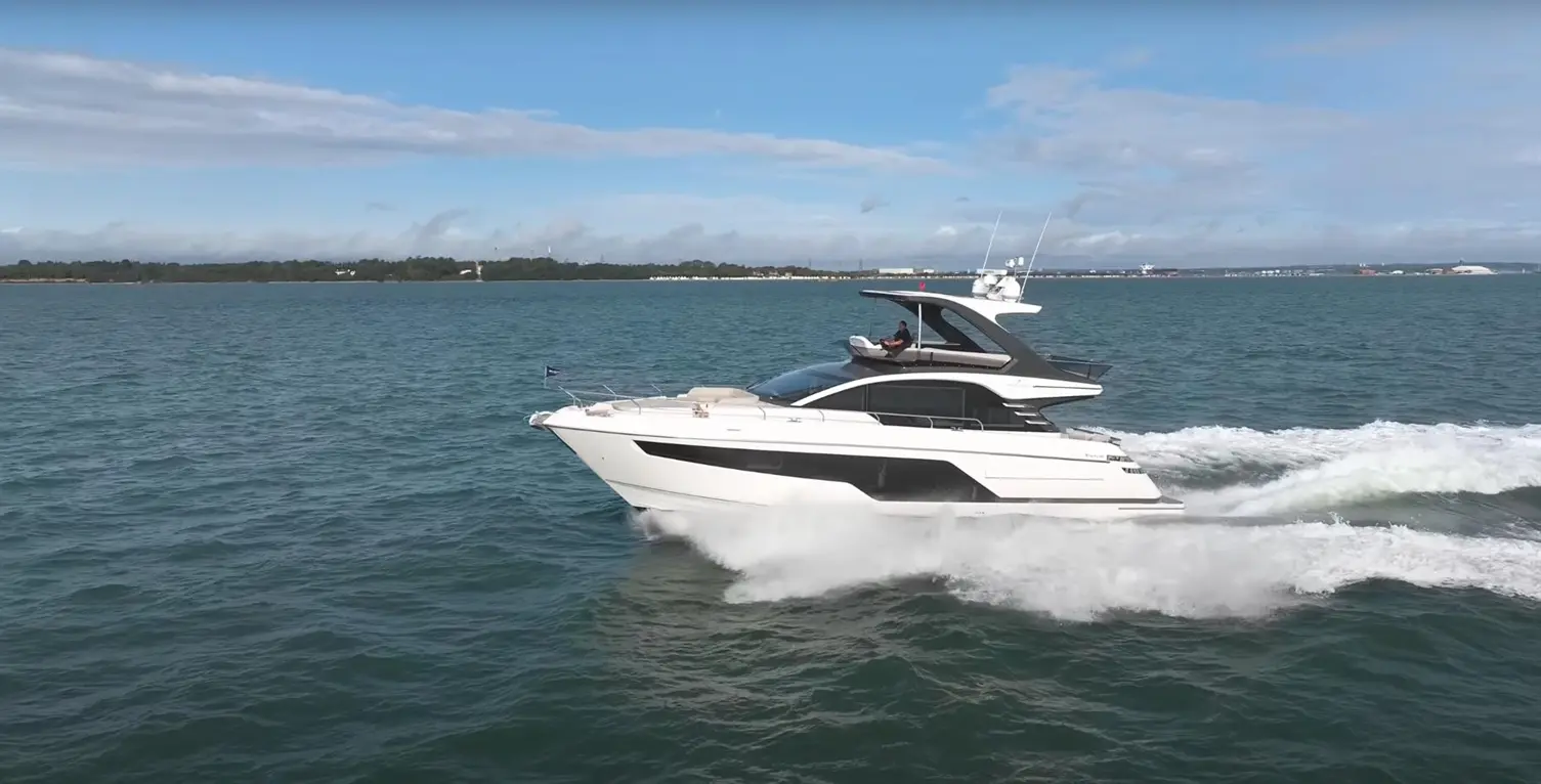 Luxury motor yacht cruising on blue water, creating a white wake, with a clear sky and shoreline in the background.